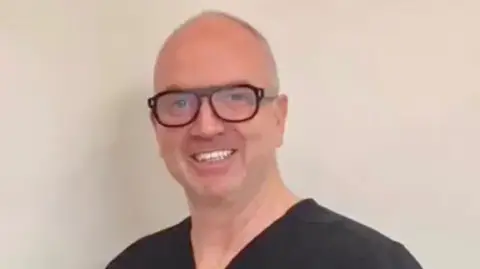 Neil Counihan is smiling into camera, in a head and shoulders photo. He's wearing black scrubs, chunky dark framed glasses and is mostly bald.