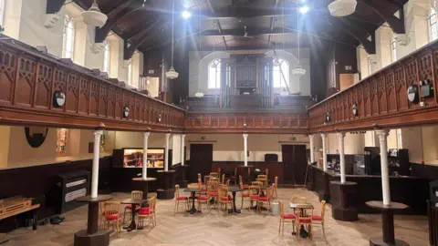 The inside of a church that has become a music venue. There is a wooden floor space with seating and small tabled, as well as standing tables around supporting beams for the balcony that runs around the perimeter of the room. 