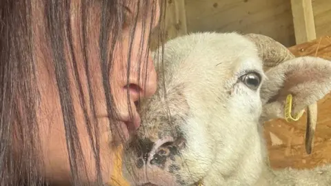 Lotus Lamb & Sheep Sanctuary A woman with dark brown hair next to a horned white sheep, which has a yellow tag on its ear.