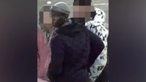 A man wearing a grey beanie and a black coat turns to look behind him. There are two people stood in front of him, one wearing a black and white coat and the other a pink coat, whose faces have been blurred.