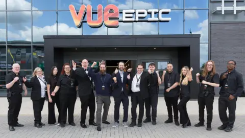 Vue Cinema Thirteen people, mostly dressed in smart black clothing, stand in a row facing the camera, standing in front of a glass cinema building, with the sign VUE EPIC fixed to it. The group are mostly cheering, with their hands and fists raised.
