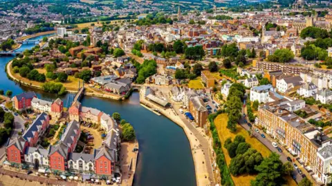 Aerial view of Exeter city. A river runs through the city. There are buildings, trees and green spaces.