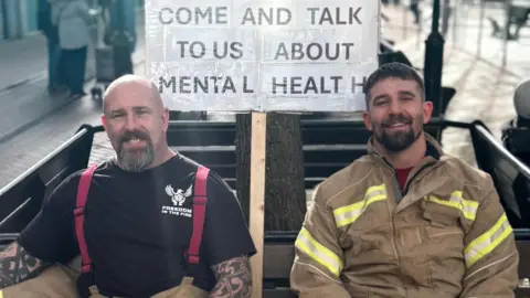 Firefighters Lee Ralph and Warren Shepherd dressed in uniform, sat on a bench. Behind them is a sign that says ' Come and talk to us about mental health'.