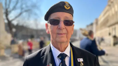 An old man with short grey hair dressed in a military suit with a military beret and a lot of military and Royal British Legion pins - including the poppy - on his blazer. He is stood on a promenade in a regency town centre on a sunny day.