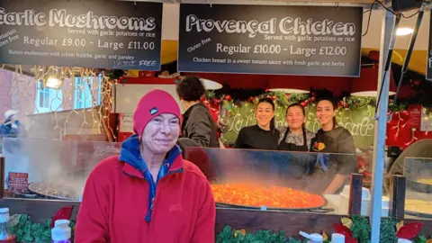 Isabelle Ecolivet, wearing a red top and beanie hat, smiles in front of her Provencal Cuisine stall with her staff smiling behind the counter as a red chicken dish simmers 