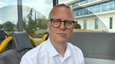 Ben Schofield/BBC Daniel Clarke on a bus. He is wearing glasses and a white shirt.