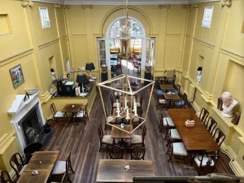 A large room with yellow walls and a dark-wood floor is viewed from above. It has a fireplace and tables laid out in small groups with chairs. Some plaster busts rest in alcoves and a metal light fitting with candle-style bulbs can be seen in the foreground. 