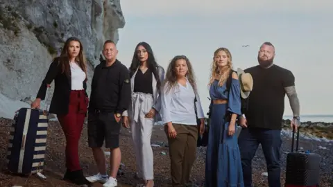 Channel 4 Six people on a beach in Dover - smartly dressed with bags and suitcases. Cliffs on the left, sea on the right, pebble beach with rocks