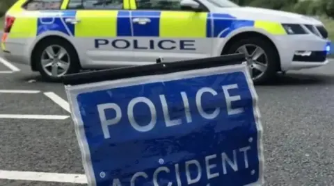 A police car parked on a road with a blue sign which reads 'police accident' positioned in the foreground.
