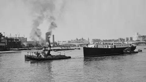 Getty Images A tugboat towing a large sloop on the river Thames in London, in black and white.