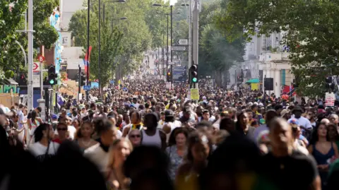 PA Media A large crowd of people fills a street during a sunny day at Notting Hill Carnival in London. The road is lined with trees and buildings, with various signs, traffic lights, and carnival decorations visible. The dense crowd stretches far into the distance.
