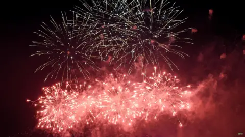 A firework display at night. There are white fireworks at the top, and pink ones below lighting up the school.
