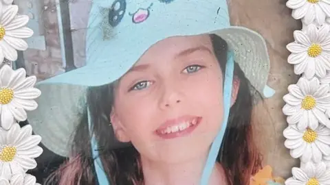 A picture in a frame. The frame is made up of plastic white daisies. The picture is of a smiling young girl with long brown hair. She is wearing a bright blue sun hat.