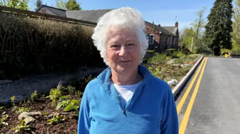 Margaret Gartside, a woman wearing a blue fleece top, is standing at the side of a road with double yellow lines along it. Next to her is a planted flowerbed. There are trees and buildings in the distance behind her.