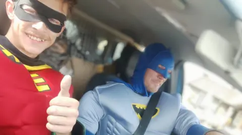 Liam Keen Two men dressed as Batman and Robin in van. Robin is giving a 'thumbs up' gesture.  