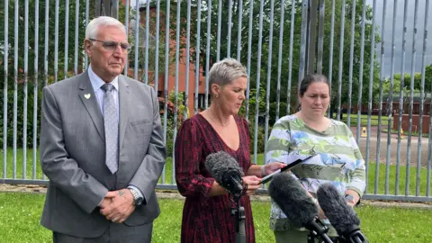 Det Ch Insp Hazel Miller alongside David and Heather Peck. Hazel has short blonde hair, wearing a long black and red dress holding a clipboard. David has white hair and wearing a grey suit. Heather has brown hair tied back, wearing a green and white striped long sleeve top. 