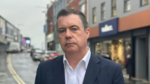 Roberts has short dark hair and is wearing a patterned suit jacket and white shirt.