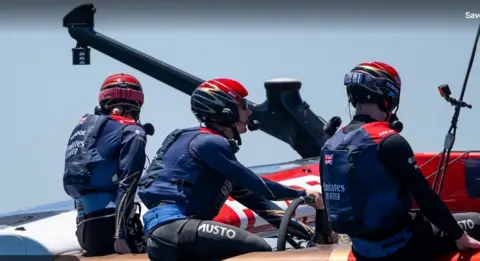 GB1 Three people in blue and black wet suits sit in a boat looking at the sea. 