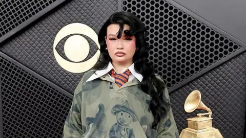 Getty Images Lola has long black hair, she is wearing a dark green jumper with a large white shirt collar over the top. She has a blue and orange striped tie on. 