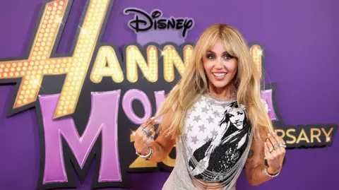 Getty Images Miley is wearing a black and white Hannah Montana t-shirt with a silver sparkly dress on top. She has long blond hair. She is stood in front of a purple background with the Hannah Montana logo in gold and purple letters. 