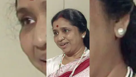 A woman, Asha Bhosle, with dark hair smiling. She is wearing a white, red and pink top.