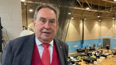 Stroud MP David Drew. He is wearing a grey suit jacket with a blue and white shirt, a red tie and red jumper underneath. He is stood on a balcony above a local election count. 