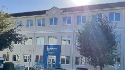A three‑storey modern building with a sign reading “Highlands College - Student Centre & Reception” above the main entrance. Several cars are parked in the spaces out front, and the sun is shining brightly above the roofline. Trees frame parts of the building, and an ambulance is visible to the left.