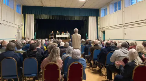 BBC People sat in rows in a community hall with their backs to the camera, and facing a stage on which four people sit behind a table