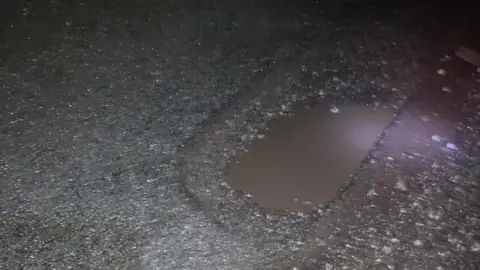 Sarah O'Connor A close-up of a hole in a road. There is some water in the hole. It appears to be night-time.
