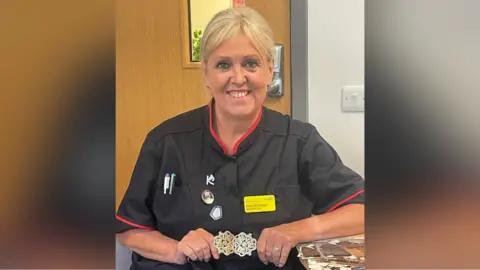 North West Anglia NHS Foundation Trust  Nurse Mary Donaldson pictured in the hospital wearing a black uniform with red lined around the sleeves and collar. She has a purple and green pen in the breast pocket of the scrubs, yellow name tag, blonde hair tied back and is smiling at the camera. She is also holding a heirloom gold and silver buckle in her hands.