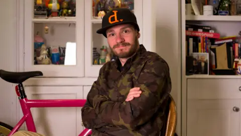 Gav Strange sat with his arms folded wearing a camo jacket and black cap. He is sat in front of white cabinets