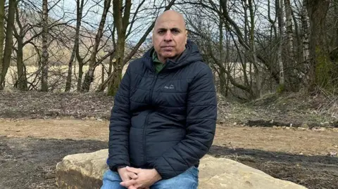 A man sits on a large rock in front of trees. He is wearing a black padded coat and blue jeans, his arms are clasped together and he has an emotionless expression on his face.