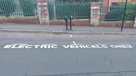 Submitted Two electric vehicle bays at the side of a hilly street. The road marking denoting their use is spelt incorrectly, reading "electric vehicels only".