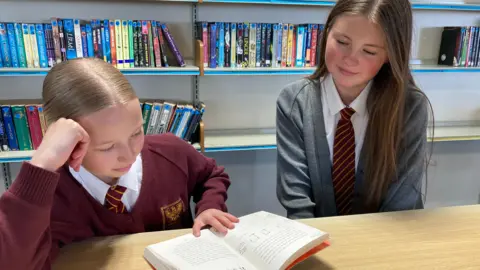 Olivia reads a book placed in the school library as Kaci-Lee sits next to her. Olivia is wearing a maroon school jumper while Kaci-Lee is wearing a grey cardigan. Both girls are also wearing maroon school ties.