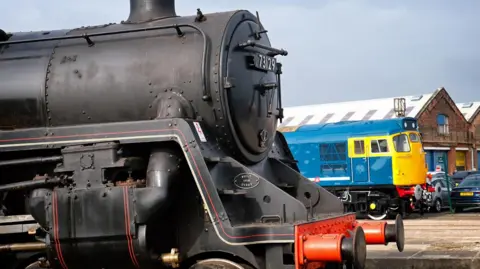 Ian Castledine A black steam locomotive is parked in the foreground, with a blue and yellow train and brick industrial buildings visible in the background 