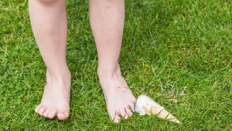 A stock image of a child who has dropped an ice cream into grass. The ice cream and their feet are visible, with the ice cream melting down the cone.