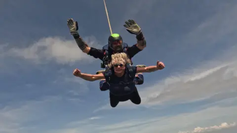 Handout Lisa skydives in tandem with an instructor. She has her arms out to her sides and thumbs up.