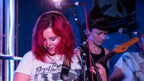 James A Mumby A woman with red hair plays guitar on stage wearing a white band T shirt for The Replacements. Behind her a woman in a black vest plays synth. She has black hair with a fringe.