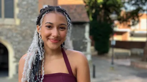Jon Wright/BBC Nandi Bushell smiling at the camera in front of a church, while wearing a purple top. She has white highlights in her hair. Behind is a stone church.