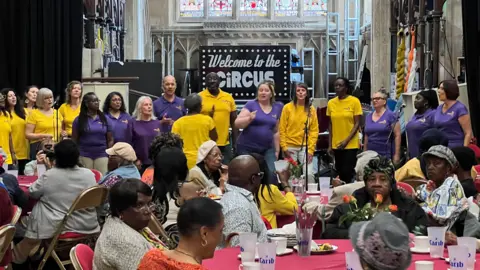 A large group of older women sit down around tables and are entertained by a group of singing people in a church.