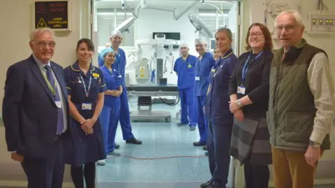 Somerset NHS Foundation Trust Staff members at Musgrove Park Hospital are standing next to each other in front of an operating room. Some are wearing blue scrubs. They are all looking at the camera and smiling.