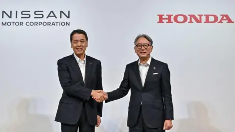 Getty Images Makoto Uchida (L), president and CEO of Japanese auto maker Nissan, shakes hands with Toshihiro Mibe (R), director, president and representative executive officer of auto maker Honda, following a press conference in Tokyo on August 1, 2024. Japanese car manufacturers Honda, Nissan and Mitsubishi Motors have signed a memorandum of understanding concerning a "strategic partnership" in the electric sector. Both men are wear blue suits with white shirts