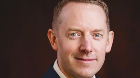 Steve Haywood Close up of Mike Badley. Mr Badley is smiling at the camera. He is wearing a white shirt and navy suit jacket. He is sitting against a dark red backdrop.