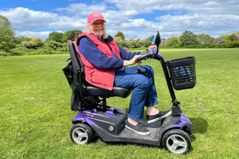 Elderley woman wearing red coat and cap whilst sat on mobility scooter in a field