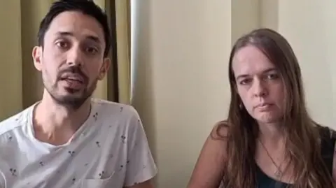 Richard Pugh and Hannah Norman sitting in a bare hotel room, looking at the camera. Richard is wearing a white T-Shirt and has short dar hair and a goatee, while Hannah has long straight hair and is wearing a dark tank top.