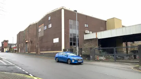 The Olympus Engineering building which stands on a site that will be developed for housing in the city. There is a big building, a gated entrance, an area under cover. A car is parked near the entrance.