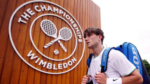 Jack Draper carries a sports bag past a sign reading The Championships Wimbledon