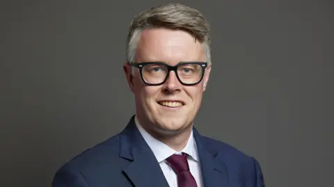 House of Commons/Laurie Noble A man with greying hair and glasses, he smiles at the camera. He is wearing a blue suit, white shirt and burgundy tie. It is an official parliamentary portrait. 