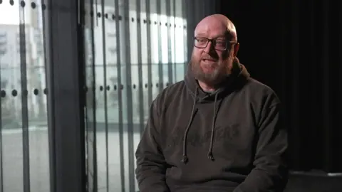 Paul Price sits in a dark hoodie, he has a beard and glasses, and appears to be in the middle of speaking to a journalist next to the camera