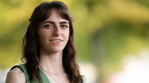 Plaid Cymru A young woman with dark brown hair, wearing a green top with a necklace looks into the camera while smiling. The background is blurred out. It is a head and shoulders shot.